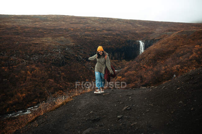 Jovem turista em óculos e chapéu com piercing olhando para baixo na colina perto de cachoeira e rio de montanha — Fotografia de Stock