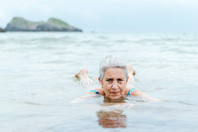 Активна старша сіра волохата жінка плаває в океанічній воді, насолоджуючись літом і практикуючи здоровий спосіб життя на морському узбережжі — стокове фото