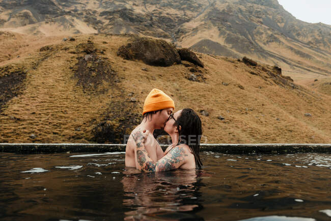 Side view of young tattooed hipsters kissing and hugging in water between mountains — Stock Photo