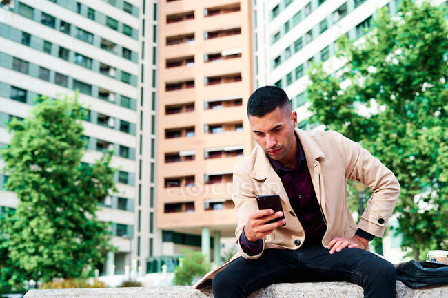 Bajo ángulo de positivo joven empresario hispano bien vestido mensajes de texto en el teléfono inteligente y discutir noticias en la calle urbana con edificios contemporáneos en el fondo - foto de stock
