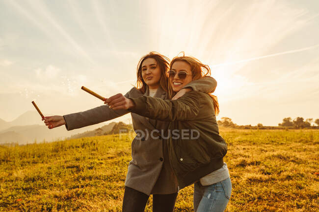 Mulheres alegres com velas cintilantes abraçando no prado nas montanhas se divertindo ao pôr do sol — Fotografia de Stock