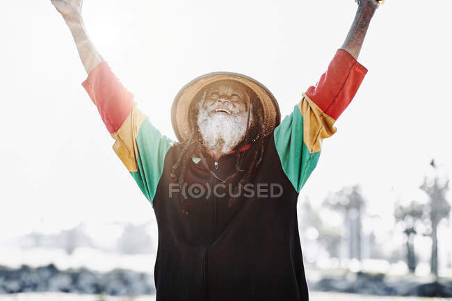 Velho e alegre rastafari étnico com dreadlocks olhando para a câmera em pé em um prado seco na natureza — Fotografia de Stock
