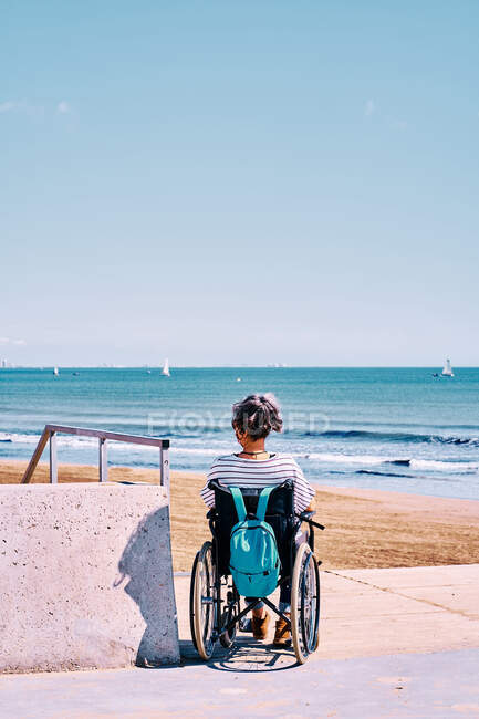 Обратный вид на неузнаваемую женщину-инвалида в защитной маске, сидящую в инвалидном кресле с рюкзаком и наслаждающуюся солнечным летним днем на песчаном пляже — стоковое фото