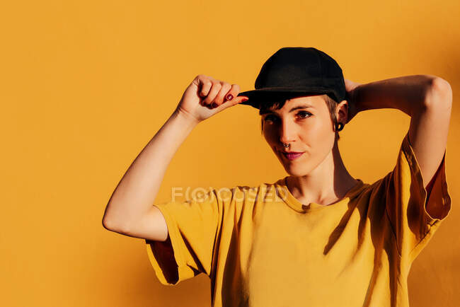 Сучасна жінка з пірсингом коригуючи модну шапку і дивлячись на жовтий фон — стокове фото