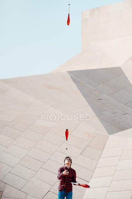 Мужчина выполняет трюк с жонглированием клубами, стоя напротив современного каменного здания с необычной геометрической архитектурой — стоковое фото
