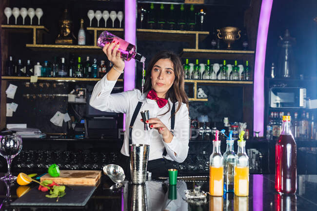Focused barista femminile in abito elegante aggiungendo liquido dalla bottiglia in jigger mentre si prepara cocktail in piedi al bancone in un bar moderno — Foto stock