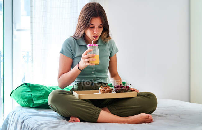 Young female enjoying healthy breakfast with fresh berries and juice while sitting with legs crossed on bed in morning — Stock Photo