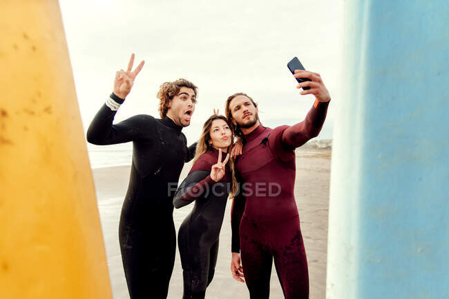 Group of happy surfer friends dressed in wetsuits standing near surfboards while taking selfie with smartphone on the beach during training — Stock Photo