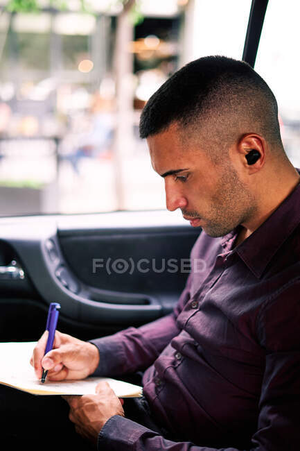 Focused Hispanic male entrepreneur sitting on passenger seat of car and writing in planner while commuting to work — Stock Photo