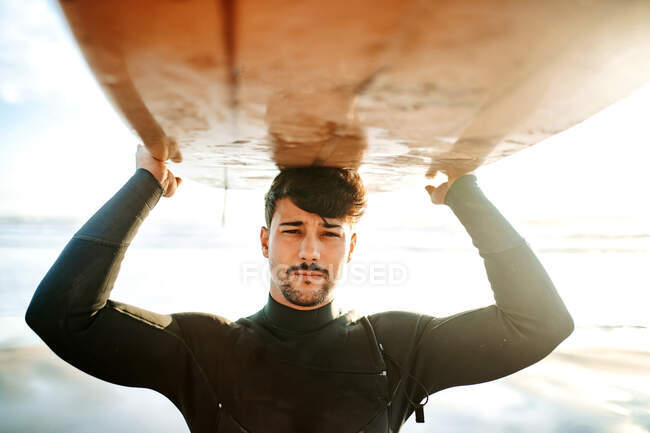 Портрет молодого серфингиста в гидрокостюме, стоящего и смотрящего в камеру на пляже с доской для серфинга над головой во время восхода солнца — стоковое фото