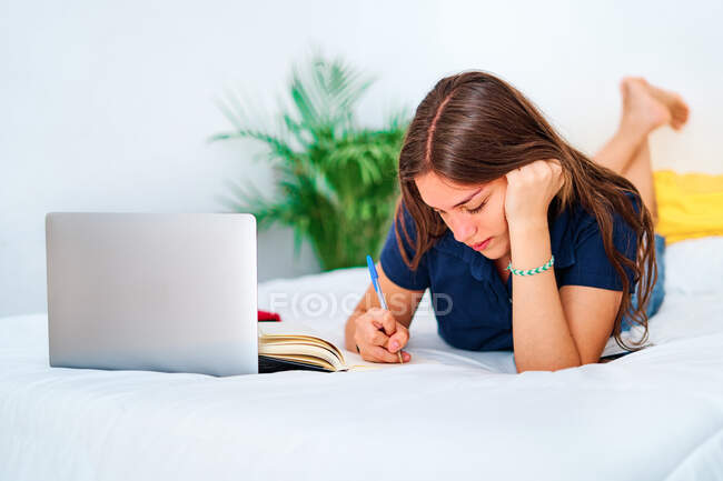 Junge Studentin liegt auf dem Bett und schreibt im Copybook, während sie während des Fernstudiums zu Hause mit Laptop online lernt — Stockfoto