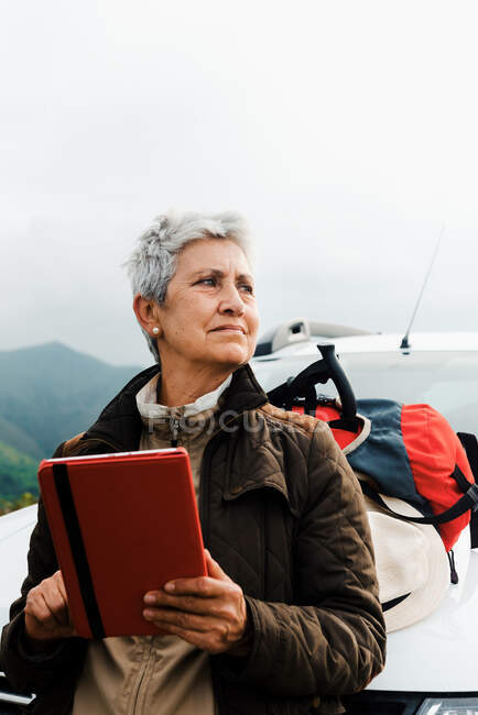 Пожилая женщина-путешественница с короткими седыми волосами, опираясь на автомобиль и просматривая планшет во время дорожной поездки в сельской местности и глядя в сторону — стоковое фото