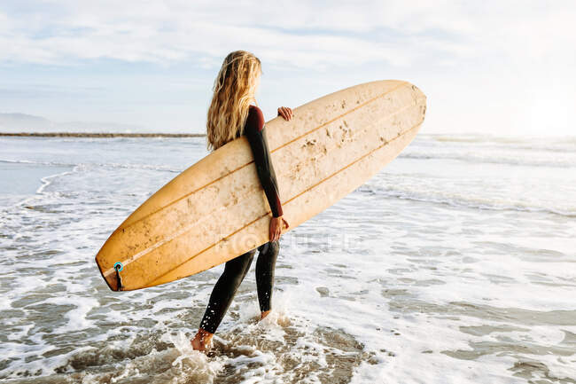 Vista lateral de la mujer surfista vestida con traje de neopreno caminando mientras lleva la tabla de surf por encima de la cabeza en la playa durante el amanecer en el fondo - foto de stock
