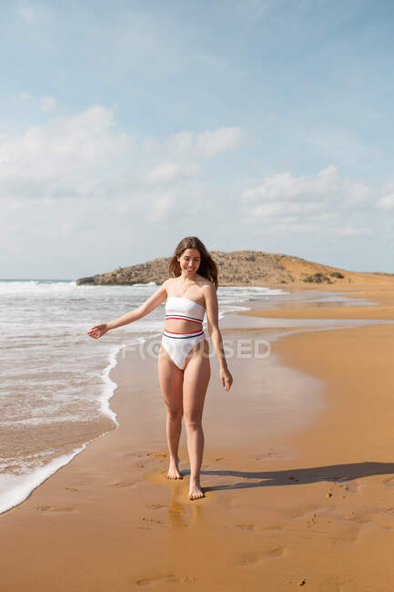 Menina sorridente em maiô em pé na praia de areia olhando para baixo perto do oceano espumoso sob o céu azul à luz do dia — Fotografia de Stock