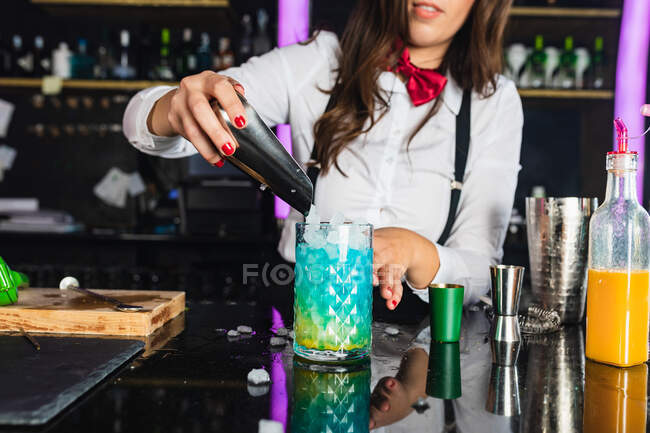 Cortado garçonete feminino irreconhecível em roupa elegante adicionando cubos de gelo em um copo enquanto prepara o coquetel azul azul em pé no balcão no bar moderno — Fotografia de Stock
