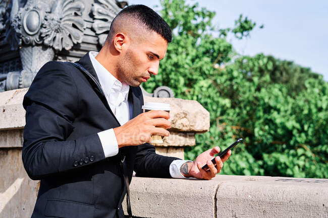 Hombre hispano de cuerpo completo con elegante atuendo mirando hacia otro lado y enviando mensajes de texto en el teléfono celular mientras se apoya en la pared en la calle de la ciudad - foto de stock
