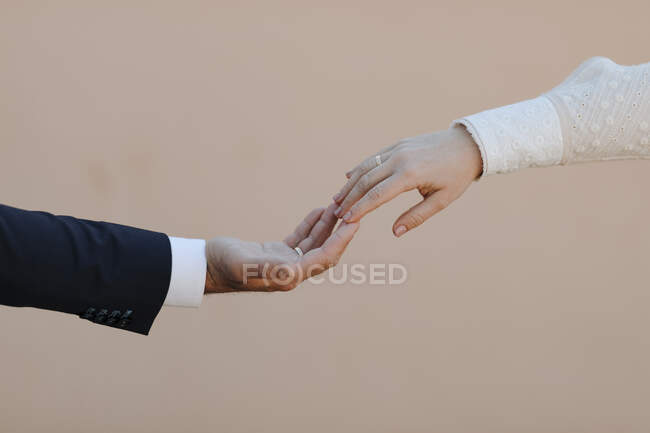 Crop casal recém-casado romântico anônimo em roupas elegantes de casamento com anéis de noivado tocando suavemente as mãos contra o fundo bege — Fotografia de Stock