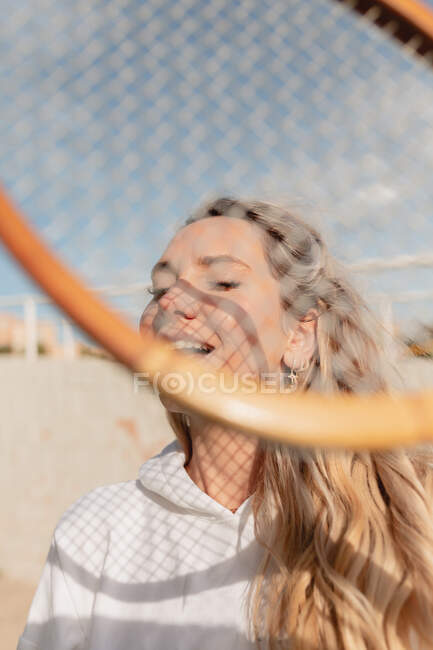 Positive junge Frau in weißen Kleidern steht mit geschlossenen Augen und Tennisschläger auf der sonnigen Straße — Stockfoto