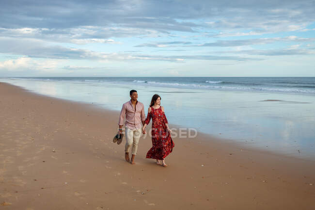 Alegre pareja multirracial cogida de la mano y caminando a lo largo de la orilla húmeda mientras admira el mar al atardecer en verano - foto de stock