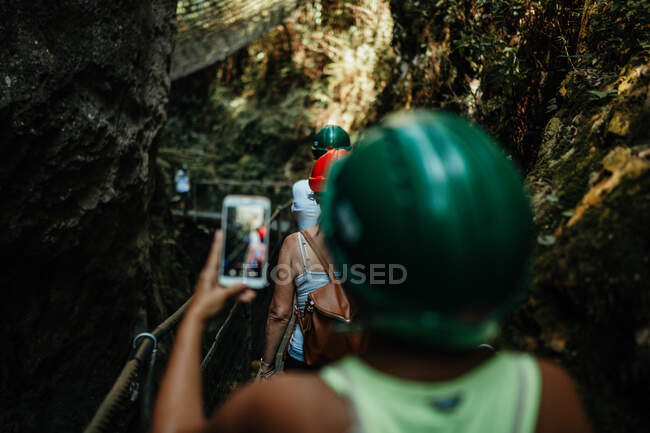 Back view of woman in safety helmet shooting on mobile phone tourists walking on footbridge between mountains — Stock Photo