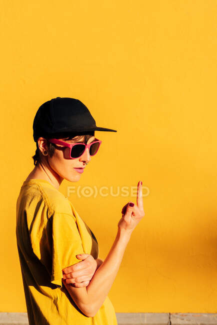 Side view of young female in street style cap and t shirt showing fuck off gesture while standing against yellow wall on city street - foto de stock
