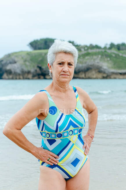 Femme âgée avec corps en forme portant un maillot de bain coloré élégant avec impression géométrique debout avec les mains à la taille contre la mer en journée d'été — Photo de stock