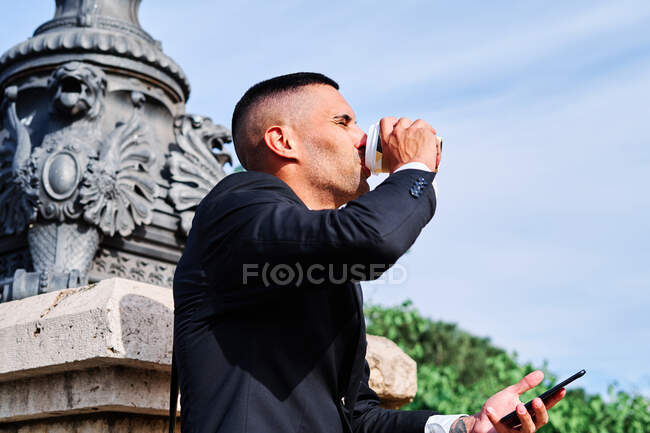Vue latérale à angle bas du jeune homme hispanique en costume noir formel avec téléphone portable à la main profitant d'une boisson rafraîchissante à emporter tout en se reposant sur la rue urbaine dans la journée d'été — Photo de stock