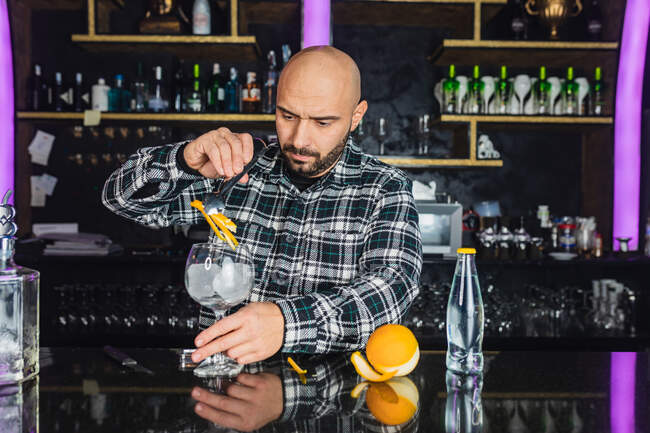 Barista maschio aggiungendo buccia d'arancia in un bicchiere mentre prepara cocktail in piedi al bancone in un bar moderno — Foto stock