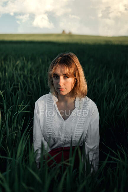 Mujer joven pacífica en blusa blanca de estilo retro sentada en medio de una hierba verde alta y mirando a la cámara mientras descansa en la noche de verano en el campo - foto de stock
