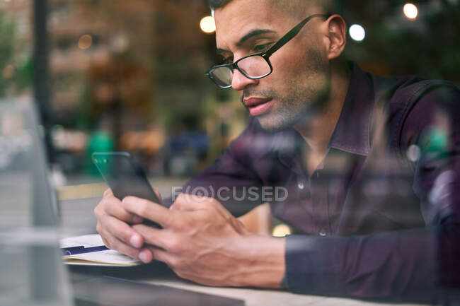 Smart Hispanic businessman in glasses making notes in notepad and answering phone call while sitting behind window in cafeteria — Stock Photo