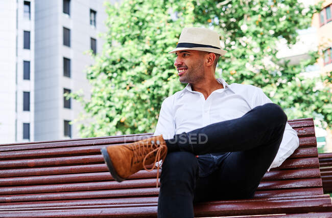 Confident handsome young Hispanic male in elegant clothes and hat sitting on bench and looking away while resting on city street — Stock Photo