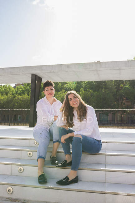 Comprimento total de jovens namoradas sorridentes em camisas brancas e jeans sentados na escada na estação perto da construção e corrimão e olhando para a câmera no dia ensolarado sob o céu azul — Fotografia de Stock
