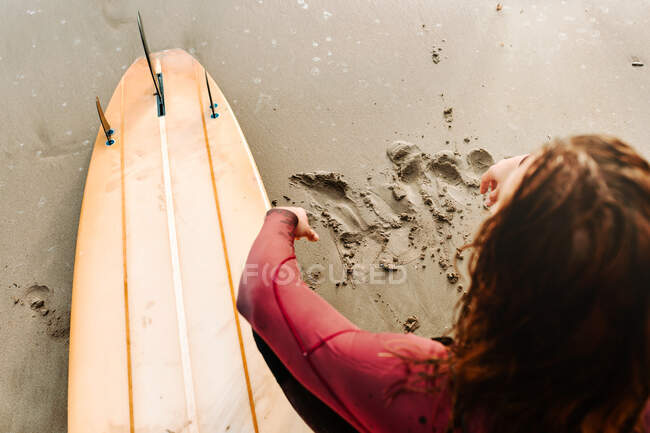 Вище невідомий серфінгіст, одягнений у вовняний костюм, сидить з серфінгом на пляжі під час сходу сонця. — стокове фото