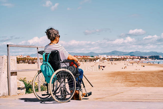 Side view of handicapped female in protective mask sitting in wheelchair with backpack and enjoying sunny summer day on sandy beach — Stock Photo