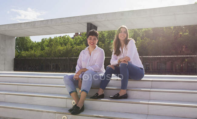 Longitud completa de jóvenes novias sonrientes en camisas blancas y vaqueros sentados en la escalera en la estación cerca de la construcción y barandilla y mirando a la cámara en un día soleado bajo el cielo azul - foto de stock
