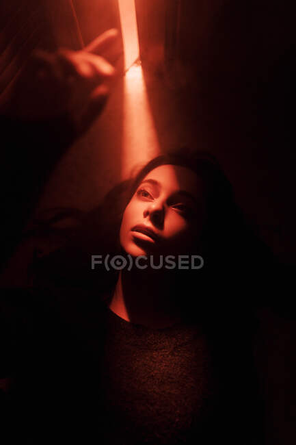Сверху тихая молодая женщина лежит на полу в темной комнате со светящимся от открытой двери светом, глядя в сторону — стоковое фото
