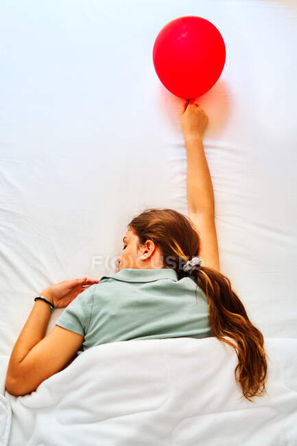 Von oben Rückenansicht einer müden jungen Frau mit rotem Luftballon in der Hand, die nach der Party mit weißen Laken im Bett schläft — Stockfoto