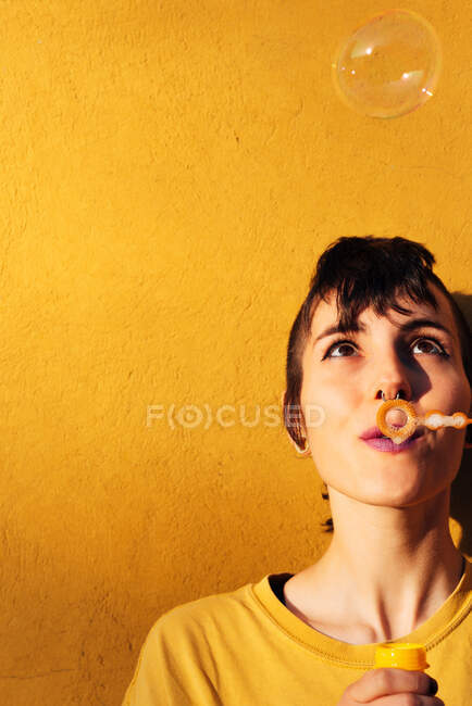 Сучасна жінка з пронизливою мильною бульбашкою на камеру в сонячний день проти жовтої стіни — стокове фото