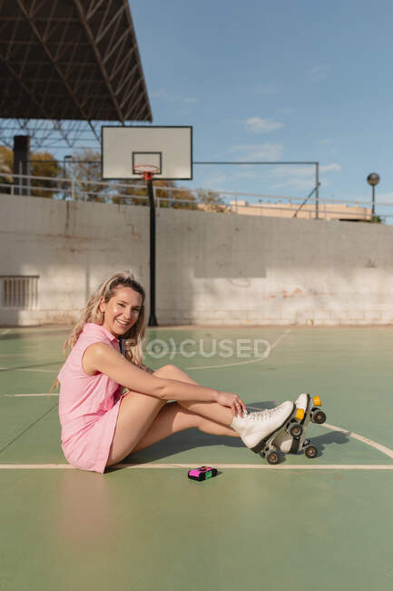 Vista laterale full length sorridente vestibilità femminile in rosa prendisole e pattini a rotelle seduti su un terreno sportivo soleggiato e guardando la fotocamera — Foto stock