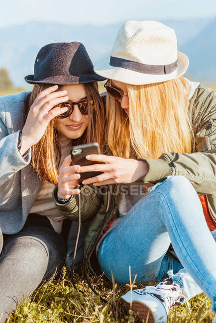Young female friends in hats and stylish clothes chilling on green lawn and sharing phone in countryside of mountains — Stock Photo