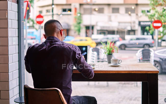 Vue arrière du mâle anonyme en vêtements décontractés intelligents assis à la table près de la fenêtre et prenant des notes tout en travaillant sur un projet à distance à la cafétéria — Photo de stock