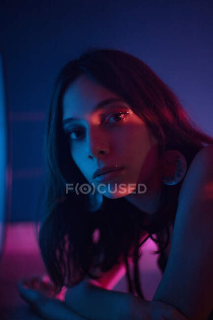 Tranquilo jovem modelo feminino no vestido deitado no chão olhando para a câmera no estúdio escuro com luzes coloridas — Fotografia de Stock