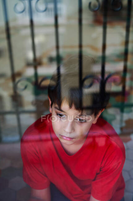 Through glass of unhappy preteen boy with bruises on face looking away while standing near window at home as concept of domestic violence and child abuse — Stock Photo