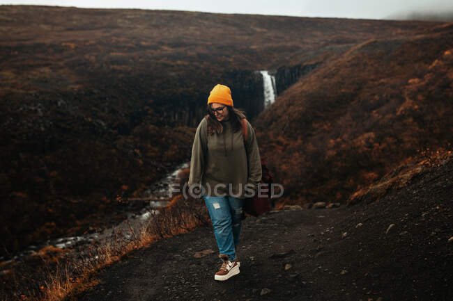 Joven turista en gafas y sombrero con piercing mirando hacia abajo en la colina cerca de la cascada y el río de montaña - foto de stock