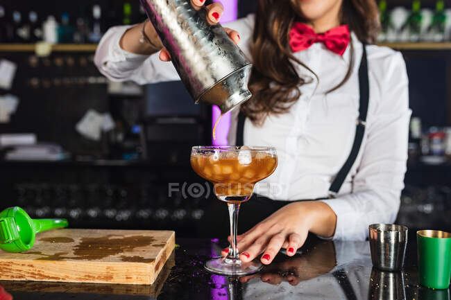 Обрезанная неузнаваемая барменша в стильном наряде, подающая коктейль из шейкера в стакан, стоя за стойкой в современном баре — стоковое фото