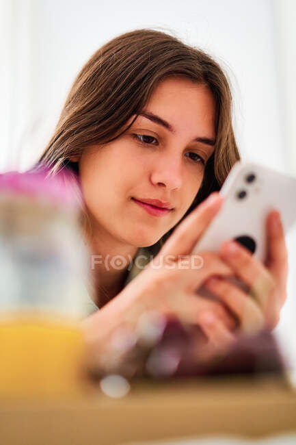 Jovem estudante navegando redes sociais no celular perto da mesa com frutas frescas e suco enquanto passa a manhã em casa — Fotografia de Stock