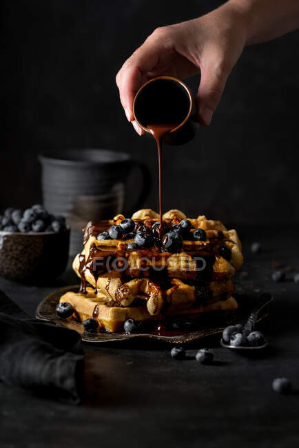 Crop anonymous person adding chocolate syrup from small cup on top of delicious waffles pyramid with blueberries on kitchen — Stock Photo