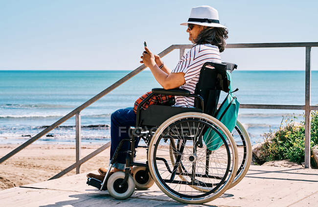 Vista lateral del viajero femenino discapacitado positivo sentado en silla de ruedas y hablando en un teléfono inteligente mientras pasa las vacaciones de verano en la playa cerca del océano ondulante - foto de stock