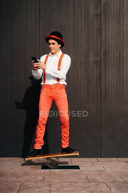 Joven artista de circo masculino navegando en el teléfono inteligente mientras está de pie en el tablero de equilibrio contra la pared gris - foto de stock