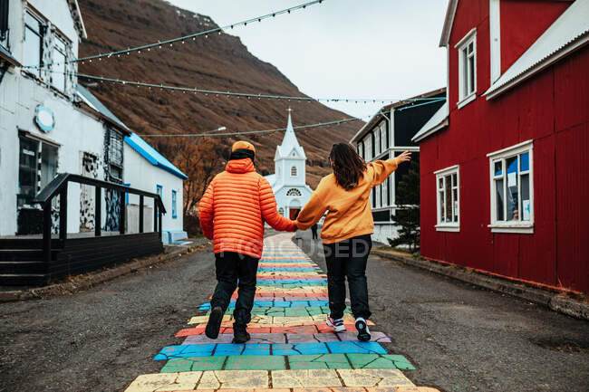 Back view of young tourists holding hands on street between buildings of village near cathedral and mountain — Stock Photo